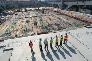 Operários de capacete e colete refletivo numa laje de obra com a ferragem armada, vistos de cima