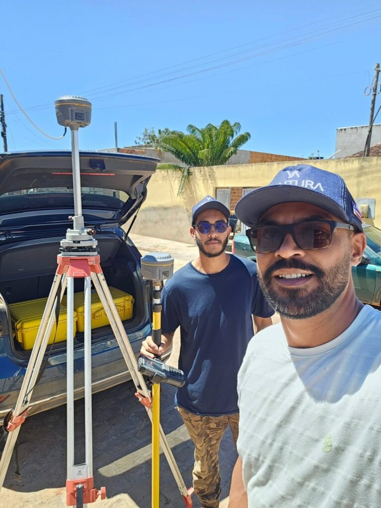 Dois técnicos da Ventura Engenharia em selfie ao lado de tripé com receptor de GPS e carro aberto
