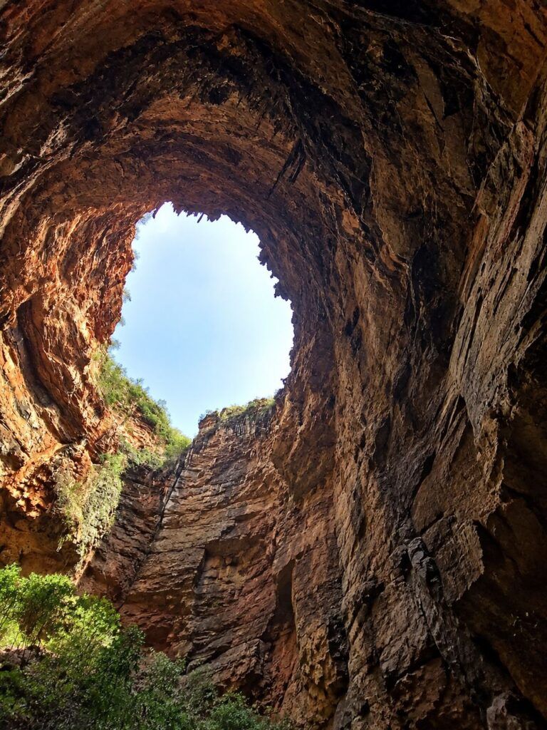 Vista de dentro da Gruta dos Brejões para uma abertura no alto, com o céu azul, paredes de rocha avermelhada e vegetação na borda