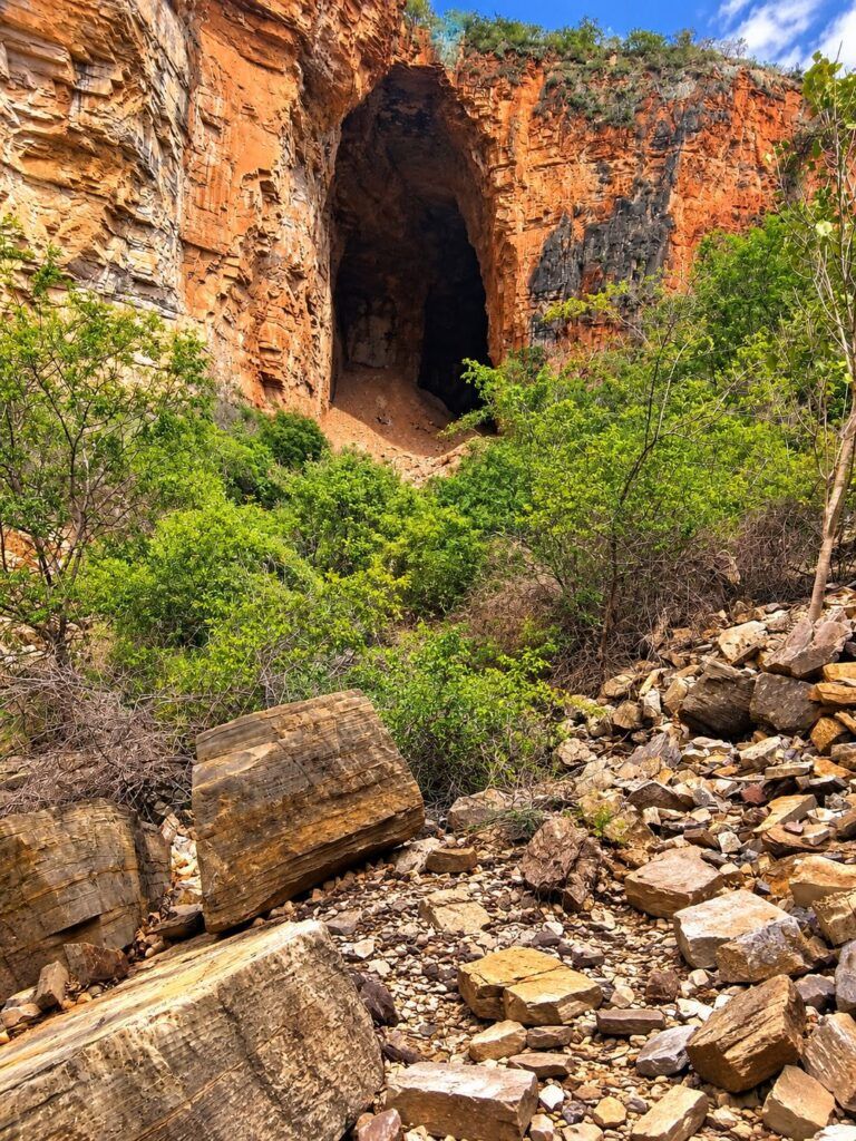 Entrada da Gruta dos Brejões aberta no paredão alaranjado, com vegetação verde e blocos de pedra na frente