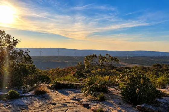 Vista do alto do Morrão, em Morro do Chapéu, com o sol baixo, vegetação e lajes de pedra em primeiro plano e aerogeradores no horizonte
