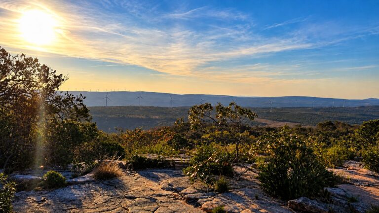 Vista do alto do Morrão, em Morro do Chapéu, com o sol baixo, vegetação e lajes de pedra em primeiro plano e aerogeradores no horizonte