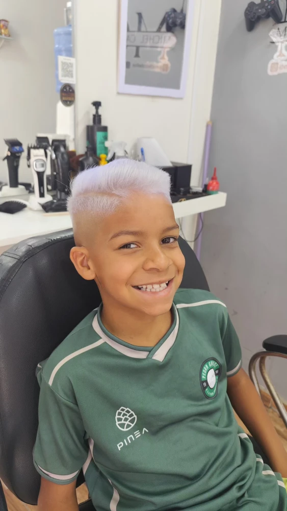Garoto sorridente sentado na cadeira da barbearia com cabelo loiro platinado