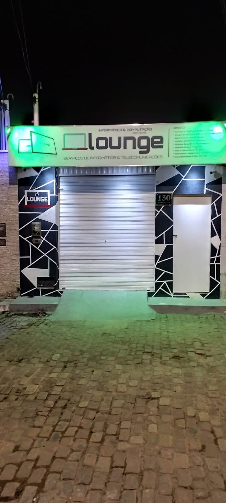 Fachada da Lounge Informática à noite, com letreiro luminoso verde e porta de enrolar, na Rua Ruy Barbosa, 130, em Morro do Chapéu