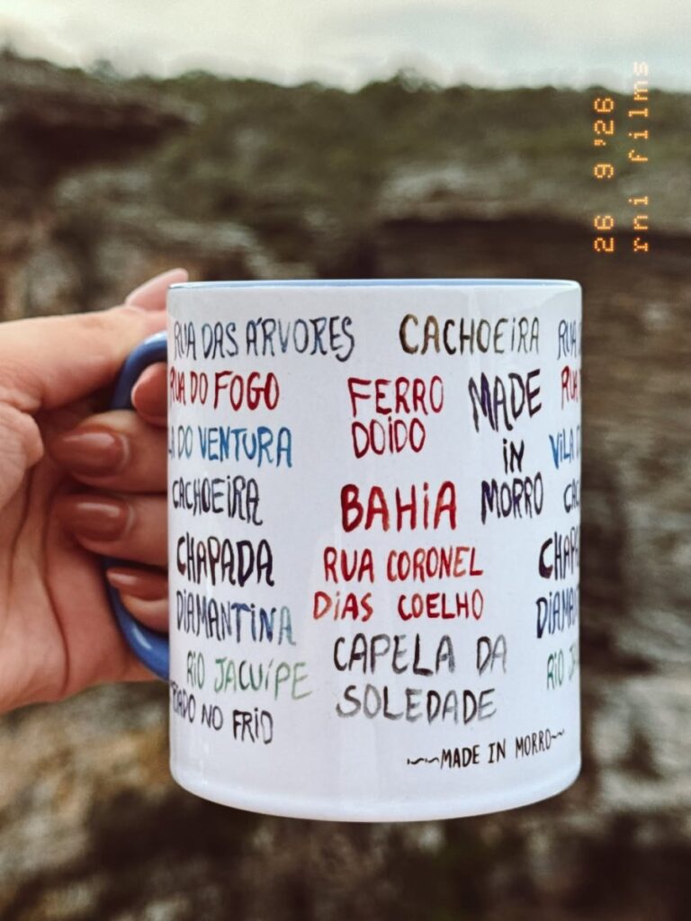 Caneca branca da Made in Morro com nomes de lugares e ruas de Morro do Chapéu, segurada diante de uma serra
