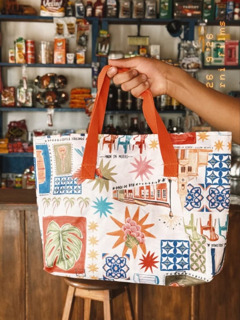 Ecobag estampada com ilustrações locais da Made in Morro, segurada em frente a prateleiras de mercado