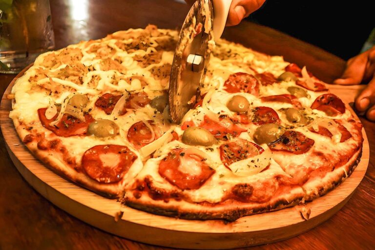 Pizza com salame e azeitonas sendo cortada por uma pessoa sobre uma tábua de madeira