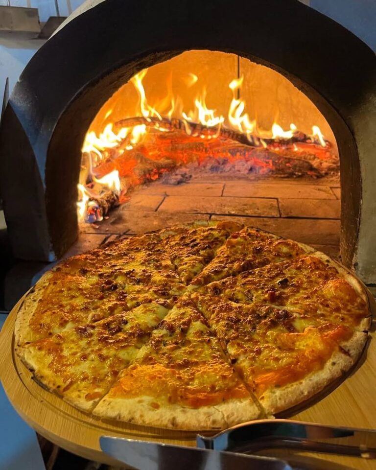 Pizza fatiada em tábua de madeira à frente de forno a lenha com chamas